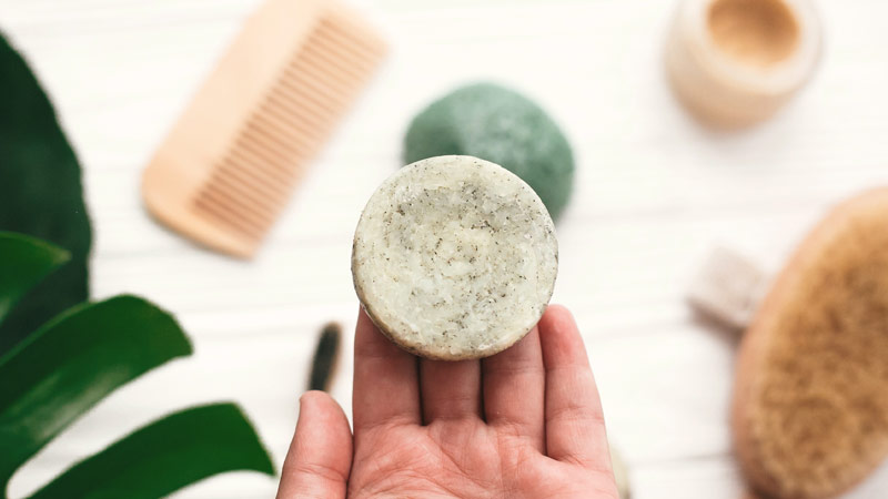 Mão de mulher com um champô sólido na mão, redondo e esverdeado, com uma bancada em fundo, onde se vê um pente, uma esponja e outros objetos de higiene pessoal